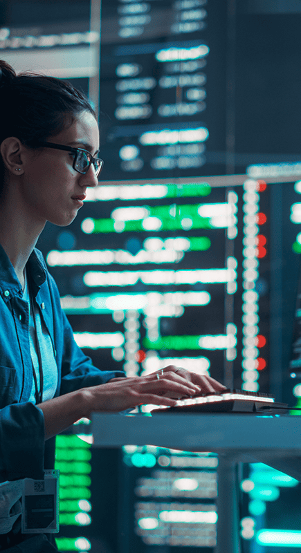 Une femme travaillant sur un ordinateur portable devant un écran d’ordinateur, fond salle de serveurs informatiques.